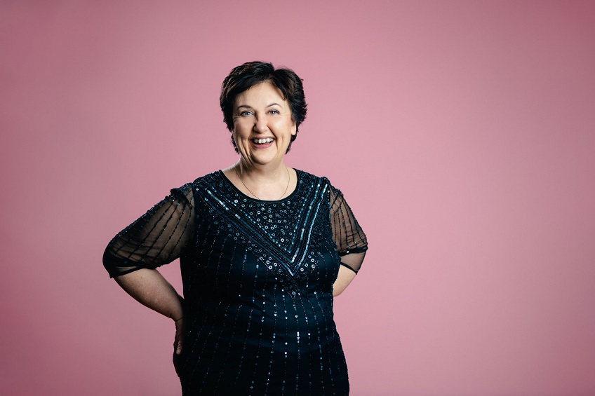 En glad Babben Larsson i svarta kläder framför en rosa bakgrund.