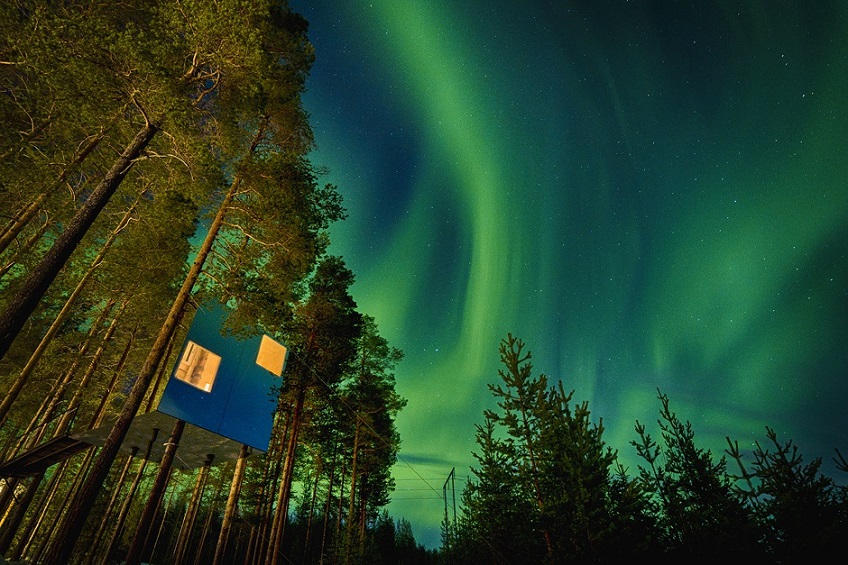 Ett fyrkantigt hotellrum som hänger i träden. Det är mörkt och norrsken ute. Det lyser från fönstren i hotellrummet.