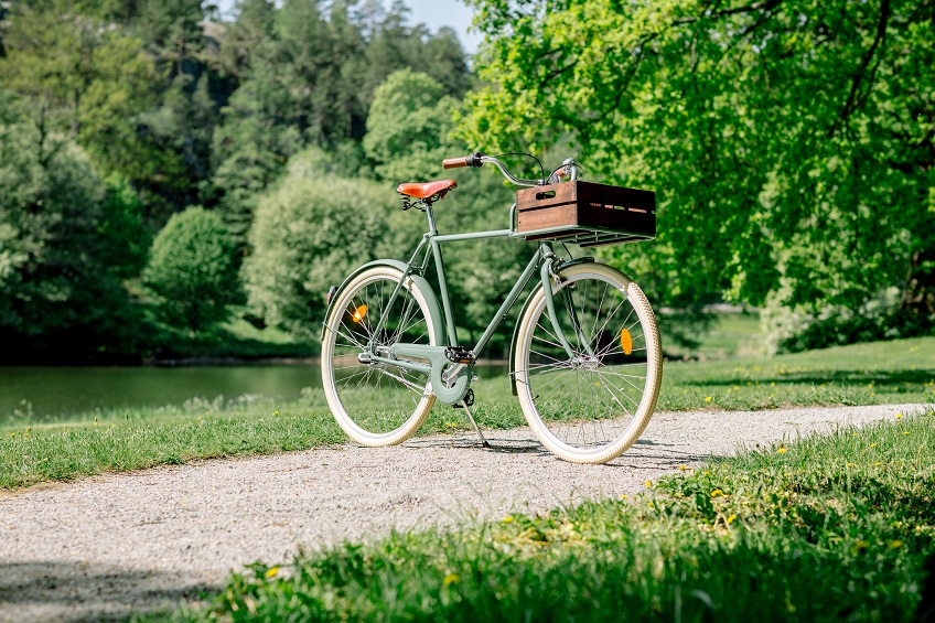 En cykel med en trälåda framför styret står uppställd på en grusgång. Grönt gräs och gröna träd i bakgrunden.