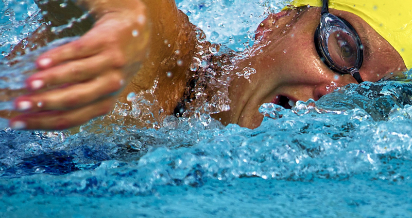 Kvinna i simglasögon och gul simmössa simmar i bassäng.